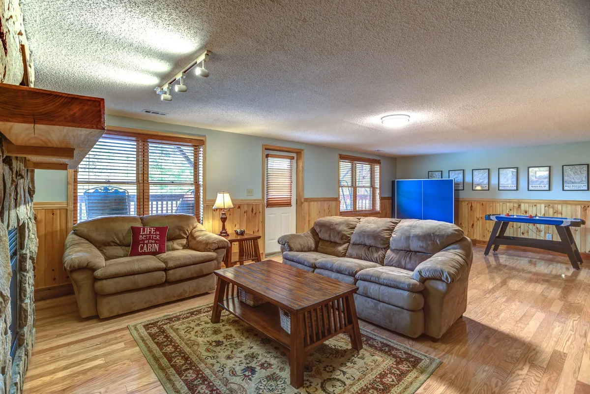 A relaxed and friendly lower-level family room with comfortable sofas, a wooden coffee table, decorative rug, and a stone fireplace. The room is arranged for gathering, playing games, and enjoying each other's company, with ample space for movement and group activities.