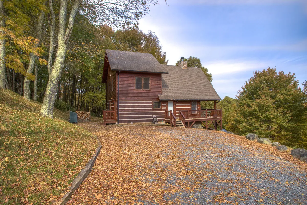 The wooden house set amidst tall trees and a carpet of autumn leaves, with a gravel driveway leading up to the porch. The surrounding landscape hints at nearby hiking and outdoor activities, while the welcoming entry showcases the property as a gathering place for families and pets alike.