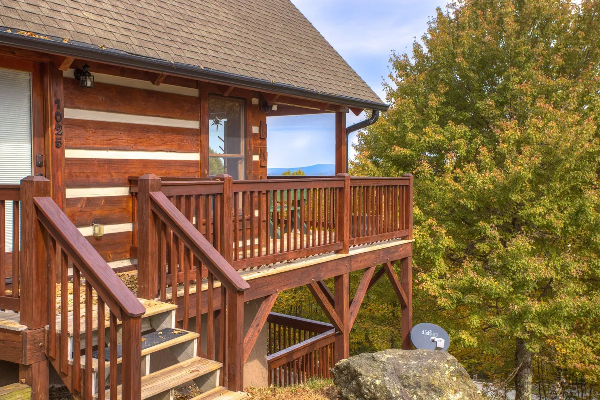 A wide shot of the log cabin structure, surrounded by lush greenery and natural mountain scenery. The wooden balcony with sturdy railings is clearly visible, inviting families to relax outdoors and take in the views. The tranquil setting emphasizes both the coziness of the home base and its connection to mountain adventures.