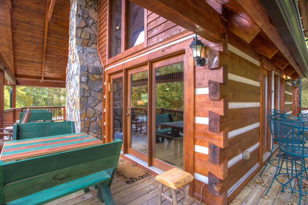 This image shows the wooden deck of the cabin, featuring outdoor seating and large glass doors that connect the interior to the natural surroundings. The cozy stone wall and log construction highlight the rustic charm, while the view offers a glimpse of the peaceful landscape beyond.