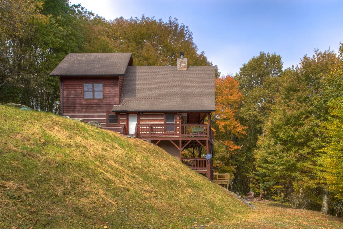 This exterior image showcases the wooden cabin nestled among trees, with a clear view of the spacious deck area that wraps around the home. The serene environment highlights opportunities for relaxing on the deck or exploring the surrounding nature.