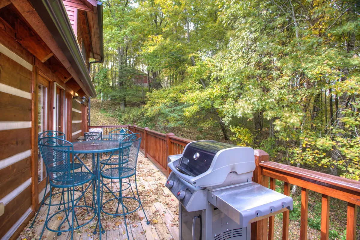 An outdoor seating area with a grill sits on a wooden deck, enclosed by a rustic railing and surrounded by lush foliage. The scene feels relaxed, perfect for gathering after a day outdoors.