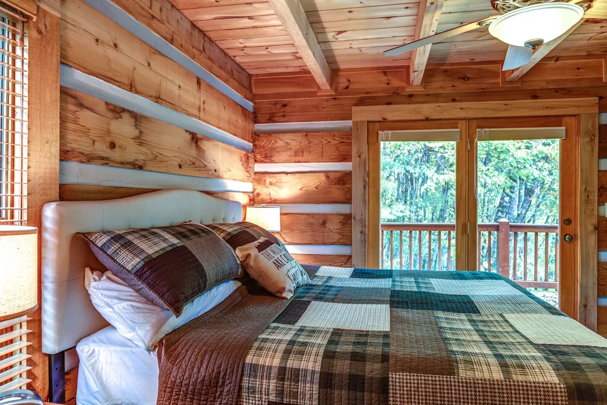 A tranquil main-level bedroom featuring a large bed with patterned bedding, wood-paneled walls, and a sliding glass door that opens onto a private balcony. Natural light pours in, creating a cozy and restful atmosphere ideal for relaxing after a day outdoors.