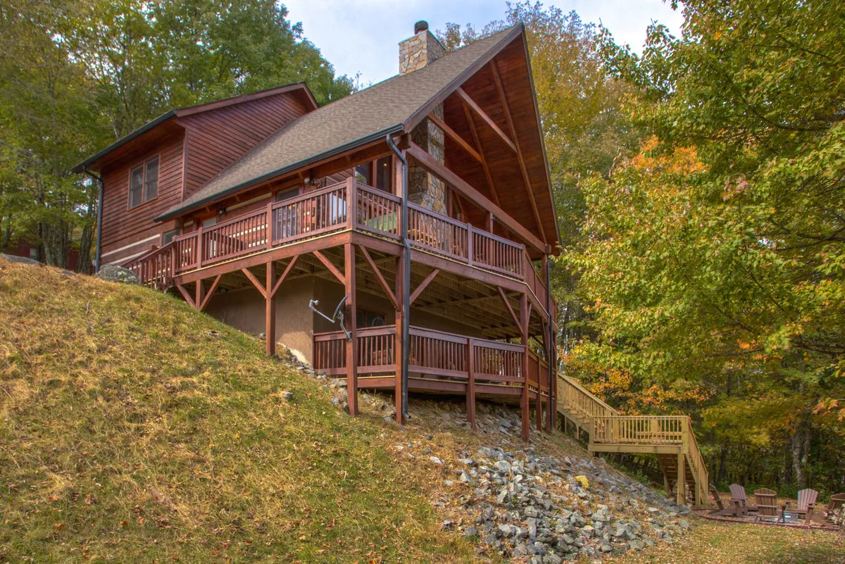 The image captures the inviting exterior of a classic log cabin, featuring a spacious wooden deck with comfortable seating, a striking stone chimney, and steps leading down to a lush natural landscape. The setting suggests the perfect place for outdoor gatherings, relaxing evenings, and taking in the surrounding mountain beauty.