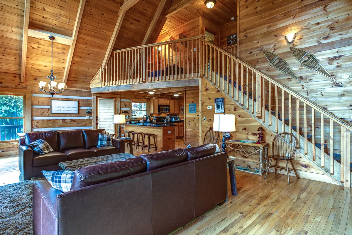 A bright, open-plan living area with a leather sofa, wooden staircase, striking chandelier, and views into the kitchen. The space feels warm and social, suggesting shared moments after a day outside. Sunlight streaming through the windows hints at a connection to the exterior deck and mountain surroundings.