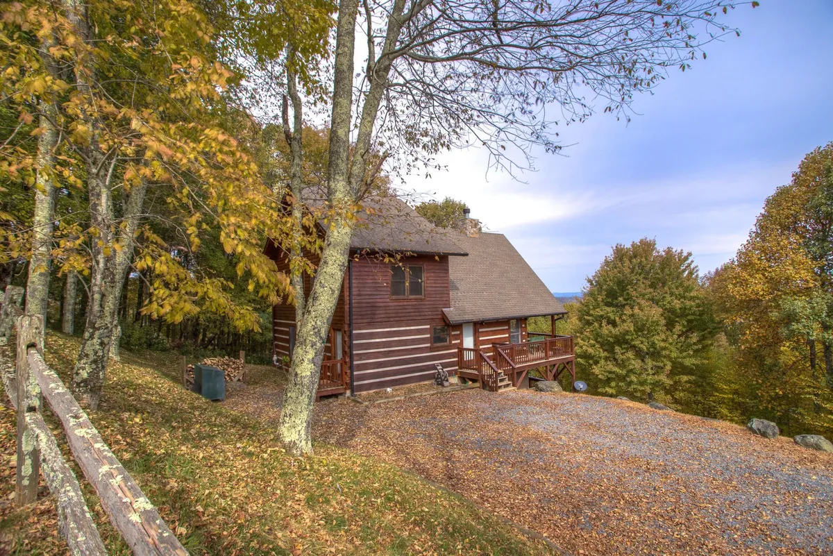 This image shows the exterior of the wooden cabin nestled among mature trees, with a gravel driveway leading up to the entrance. The inviting porch and the surrounding woods hint at both seclusion and easy access to nature.
