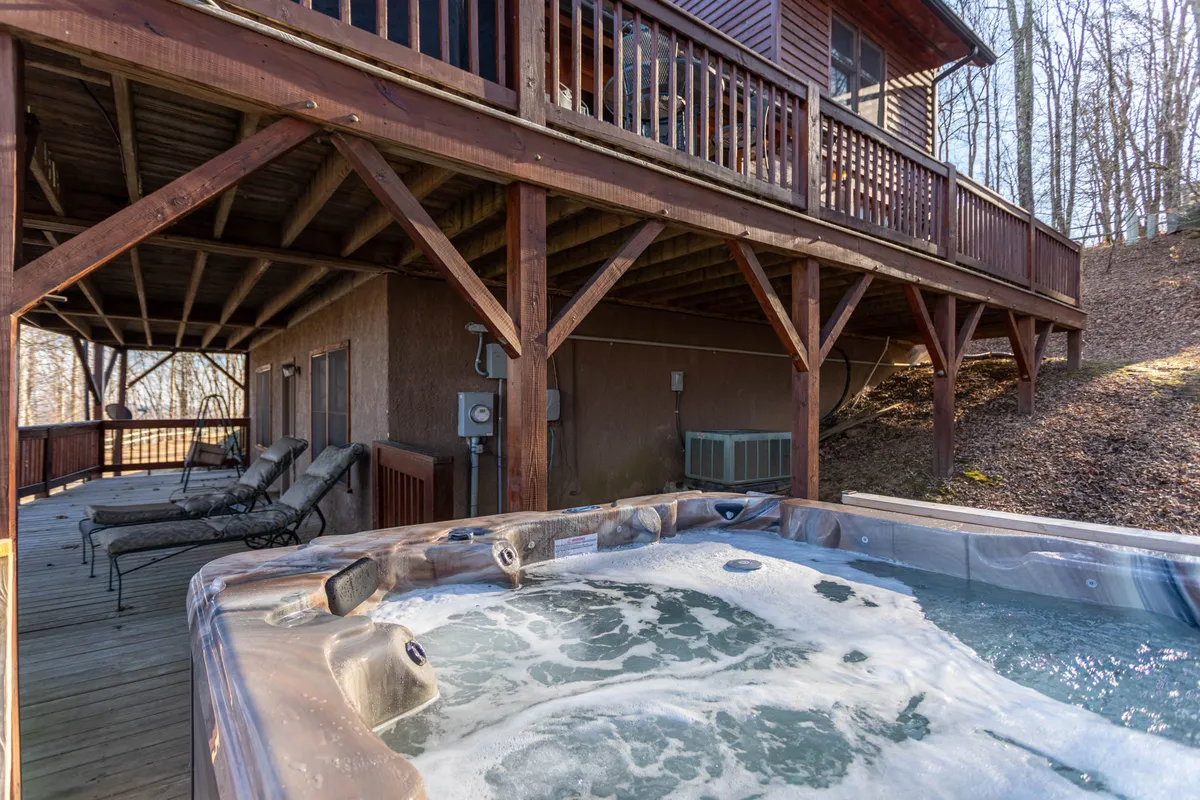 A view of the deck featuring a hot tub and comfortable reclining chairs, surrounded by wooden railings and mountain foliage. The hot tub is the focal point, directly referencing the relaxing family activities available after outdoor adventures.