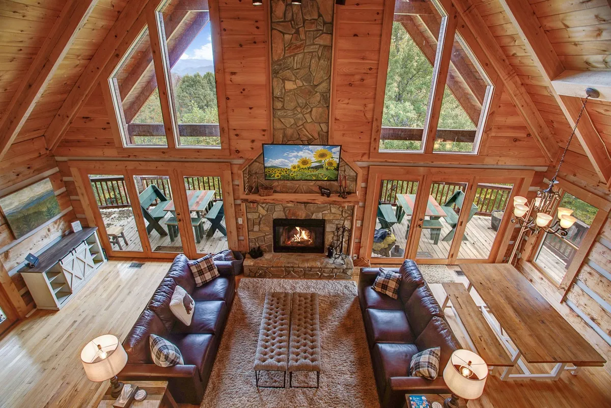 A spacious open-concept living area with a large stone fireplace as the centerpiece, plush sofa seating, a dining table, and expansive windows letting in natural light and revealing mountain views. A television sits above the fireplace, and the layout allows for seamless interaction between the living, dining, and kitchen spaces.